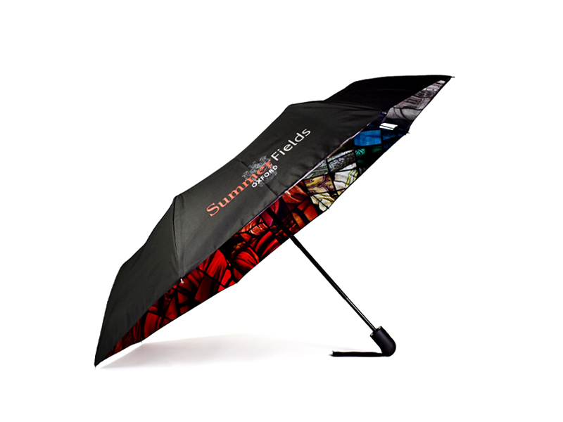 Side open view of folding umbrella. External umbrella panels are black with one panel printed with white and red text that advertises independent school Summer Fields Oxford. The inside of the panels are printed and are a stained glass window in rainbow colours.