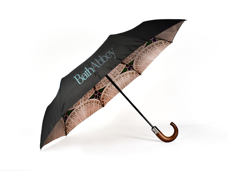 open umbrella on its side with black external panels showing with Bath Abbey text print and underside of umbrella showing with stained glass window print. The umbrella has a dark wooden curve handle