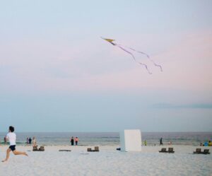 Man running with kite on a beach with pale sand with the hazy blue and pink sky behind him